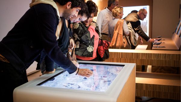 Two people exploring an image of a rock on a large display integrated in a table. The context is a museum, other people in the background.