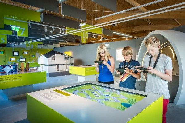 Three children in a museum next to a large display on a table. Each kids holds a device in their hands.